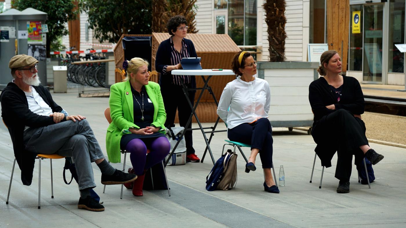 Vier Teilnehmende sitzen auf Stühlen und hören einem Vortrag zu